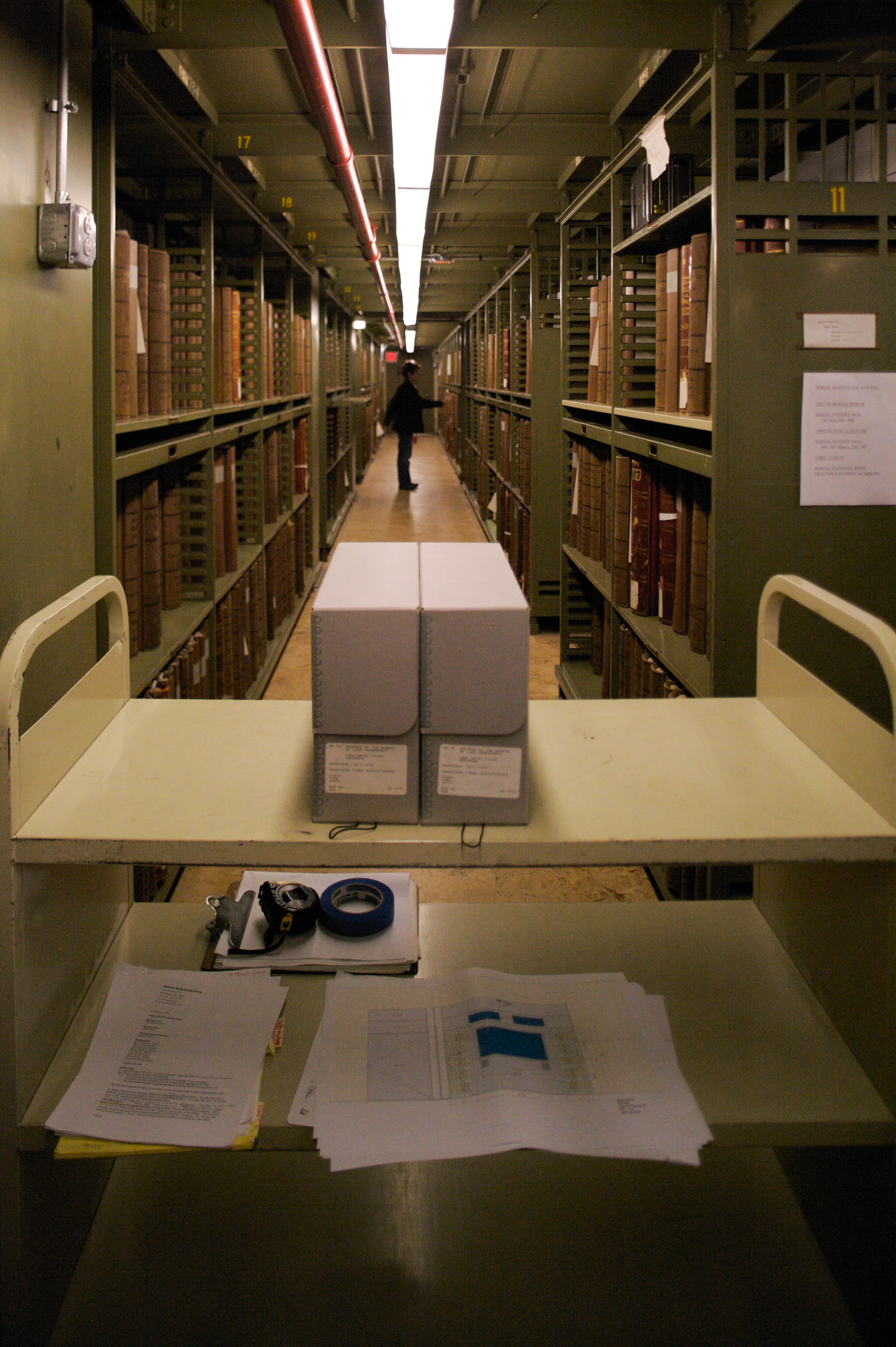 Color photograph of archival stacks. In the foreground is a cart with two archival boxes on top, in the background of the photo is a person in the stacks.